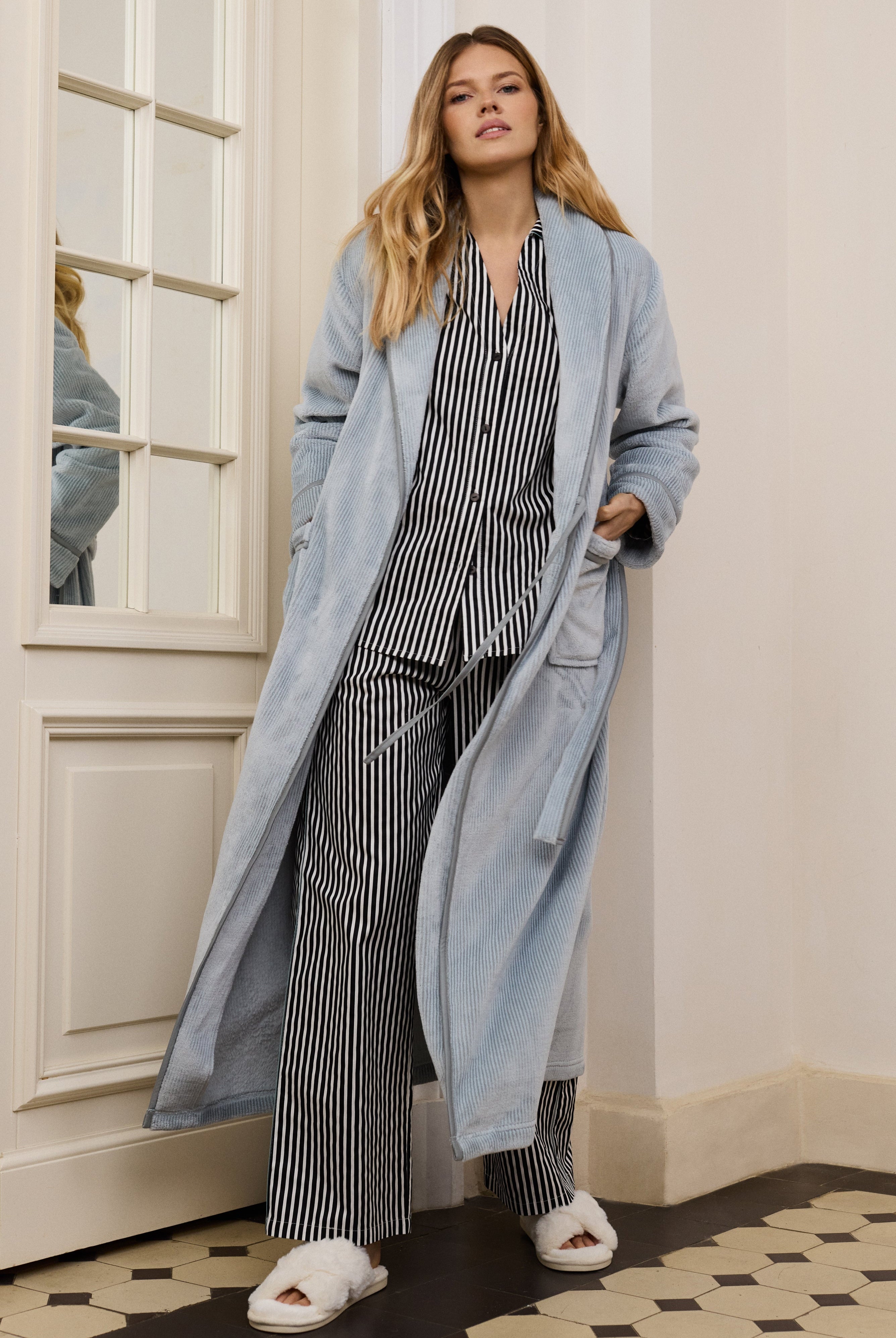 Woman wearing a long light blue bathrobe over a striped outfit in a room with white walls and a patterned floor.