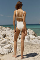 Woman in a white swimsuit standing on a rocky beach with clear blue water and sky.