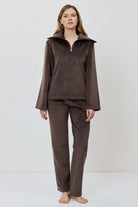 A woman modeling a matching two-piece lounge set in dark brown, featuring a long-sleeve half-zip pullover and coordinating pants.