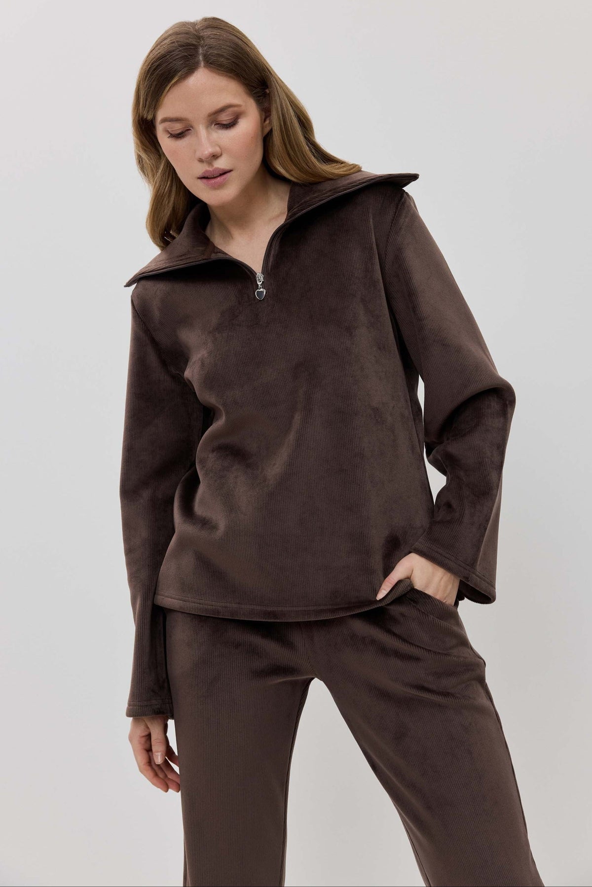A woman modeling a matching two-piece lounge set in dark brown, featuring a long-sleeve half-zip pullover and coordinating pants.