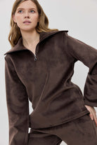 A woman modeling a matching two-piece lounge set in dark brown, featuring a long-sleeve half-zip pullover and coordinating pants.