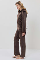 A woman modeling a matching two-piece lounge set in dark brown, featuring a long-sleeve half-zip pullover and coordinating pants.