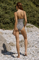 Woman in a patterned swimsuit standing on a pebbly beach with greenery in the background